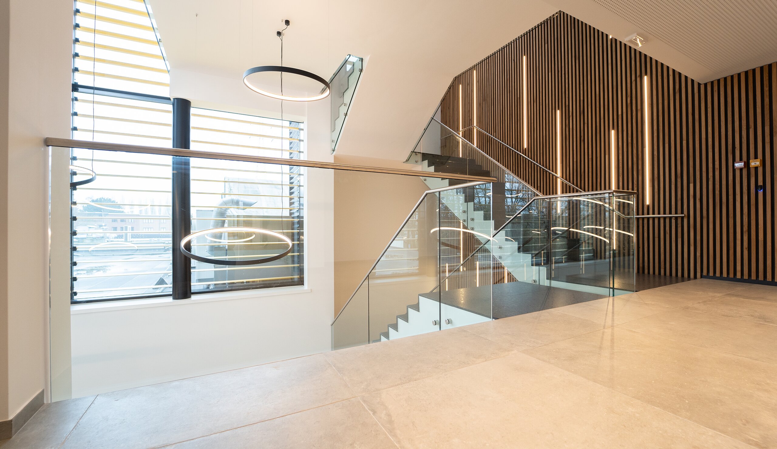 Escalier de bureaux bordé de garde-corps en verre à pinces, suspension circulaire et habillage en lattes de bois