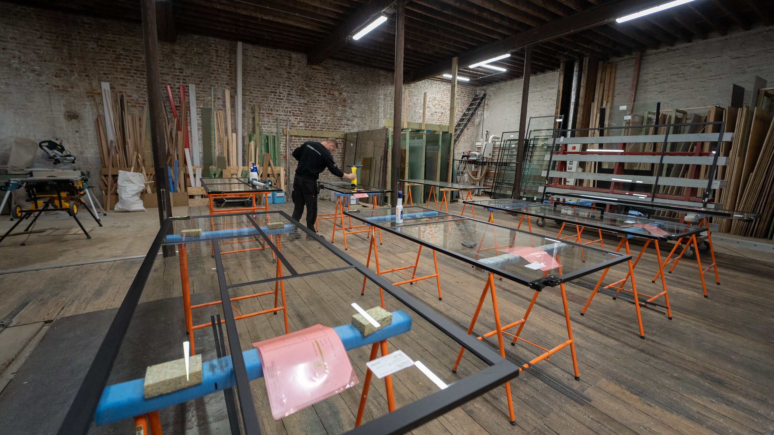 Atelier en brique et parquet ancien où sont assemblés des châssis steel look sur tréteaux orange, vue d'ensemble