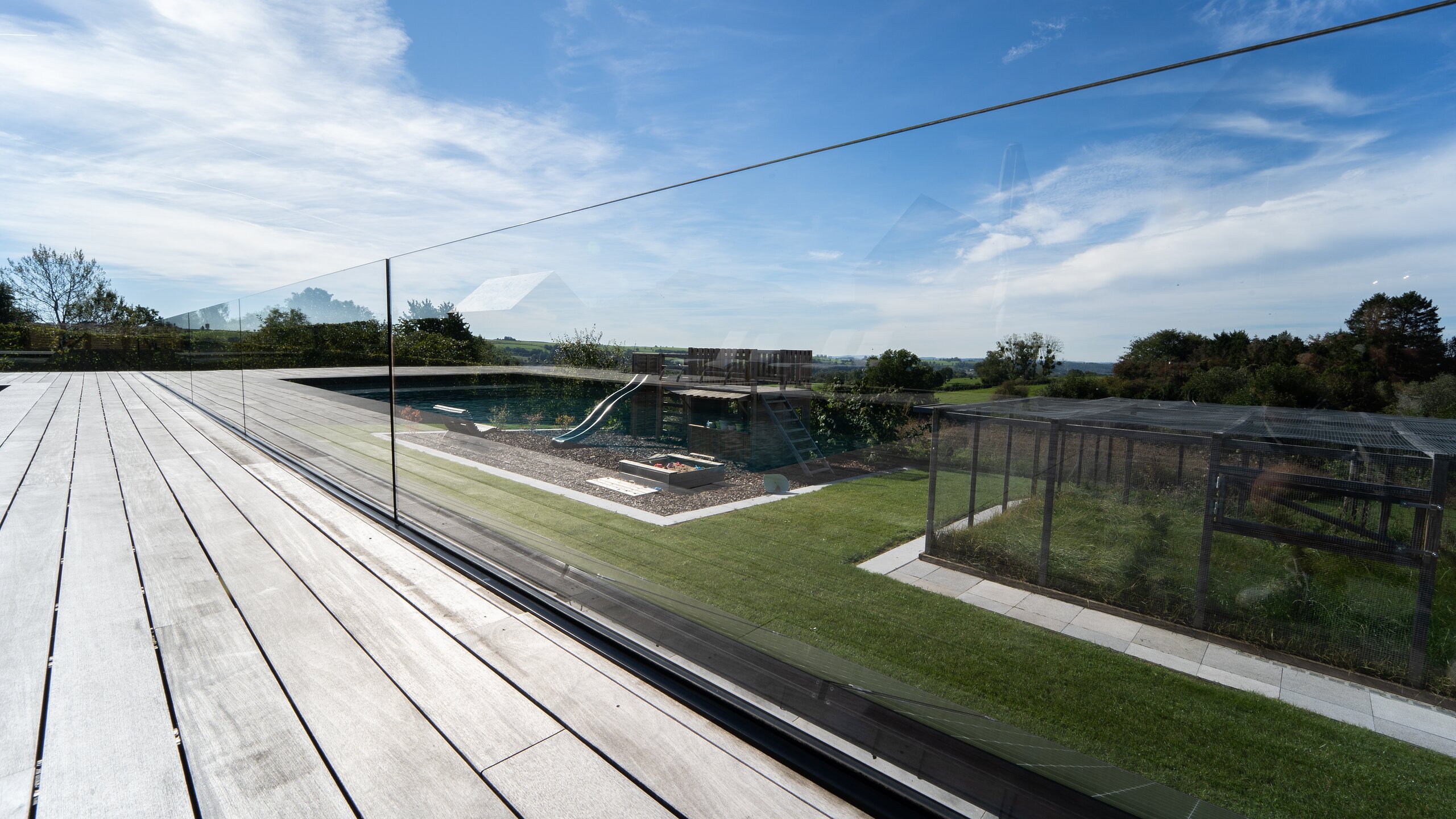 Long garde-corps en verre sans montant bordant une terrasse de piscine en bois, vue dégagée sur la campagne