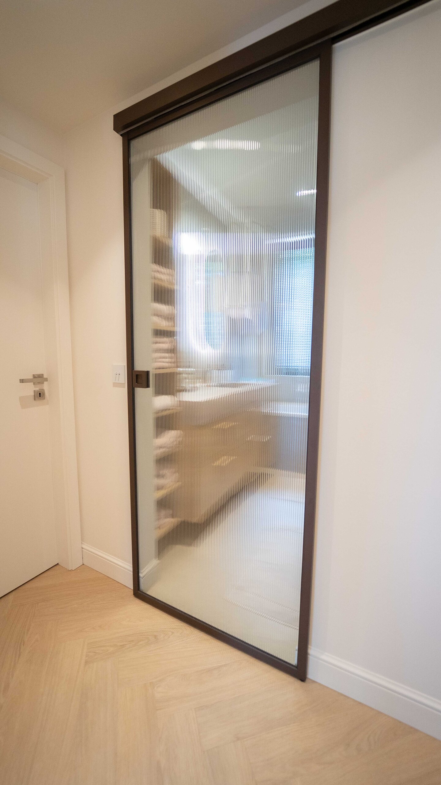 Porte coulissante en verre clair à cadre noir sur rail apparent, ouvrant sur une salle de bain aux tons bois