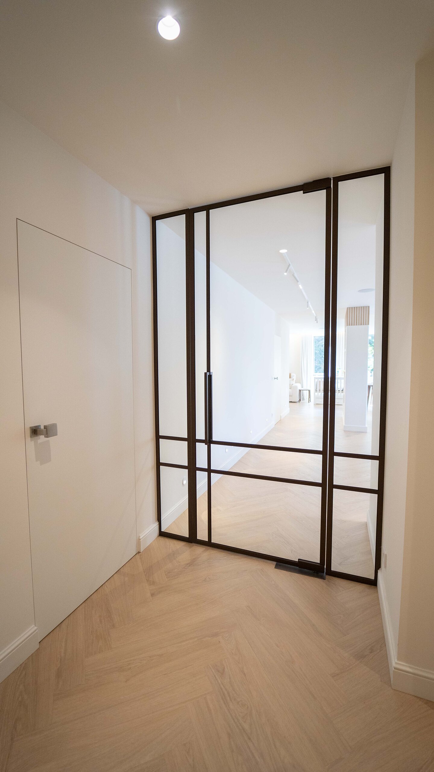 Porte pivotante vitrée à profilés noirs façon atelier avec panneau fixe, au bout d'un couloir en parquet chevron clair