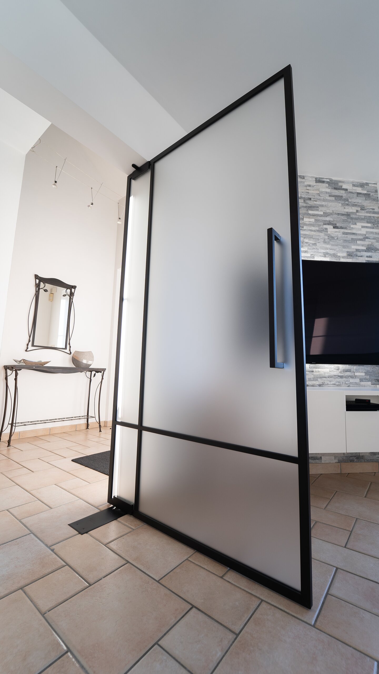 Porte pivotante en verre dépoli et traverse noire ouverte sur son axe central, sol en carrelage beige