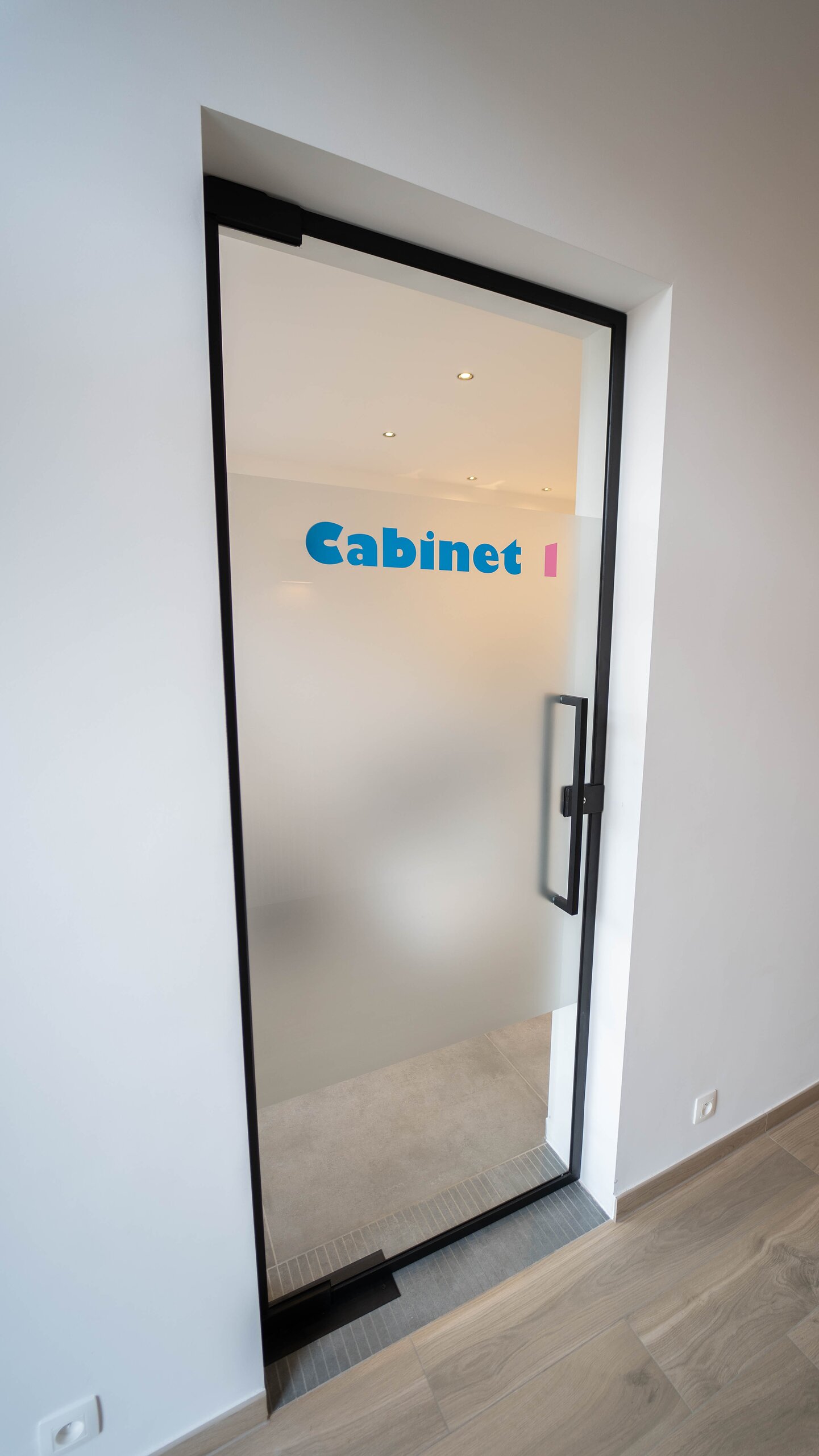 Porte en verre dépoli sur pivot à cadre noir et poignée barre, lettrage bleu Cabinet 1 appliqué sur le vitrage