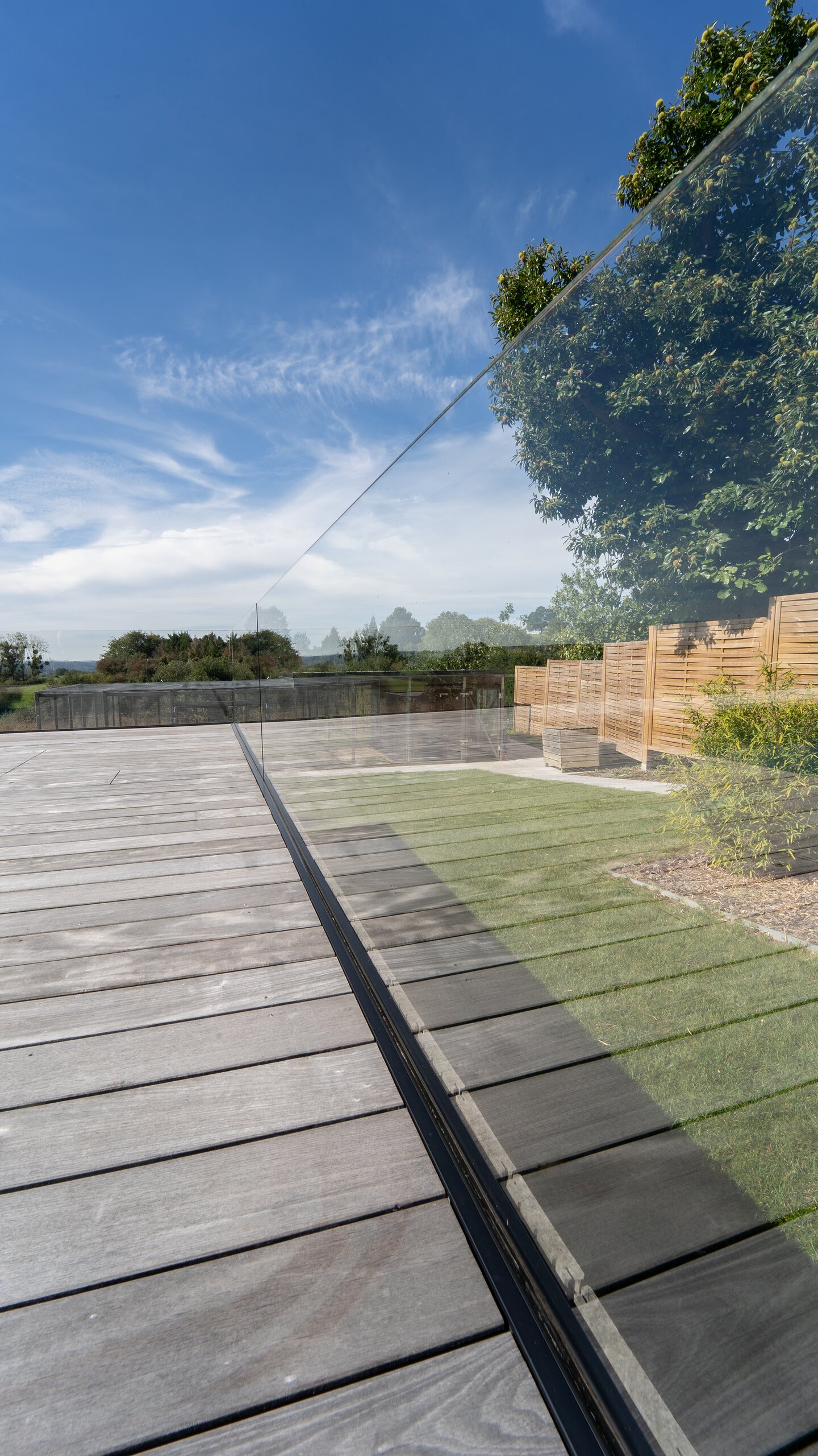 Profil au sol encastré dans une terrasse en bois, retenant un garde-corps en verre clair face à la campagne